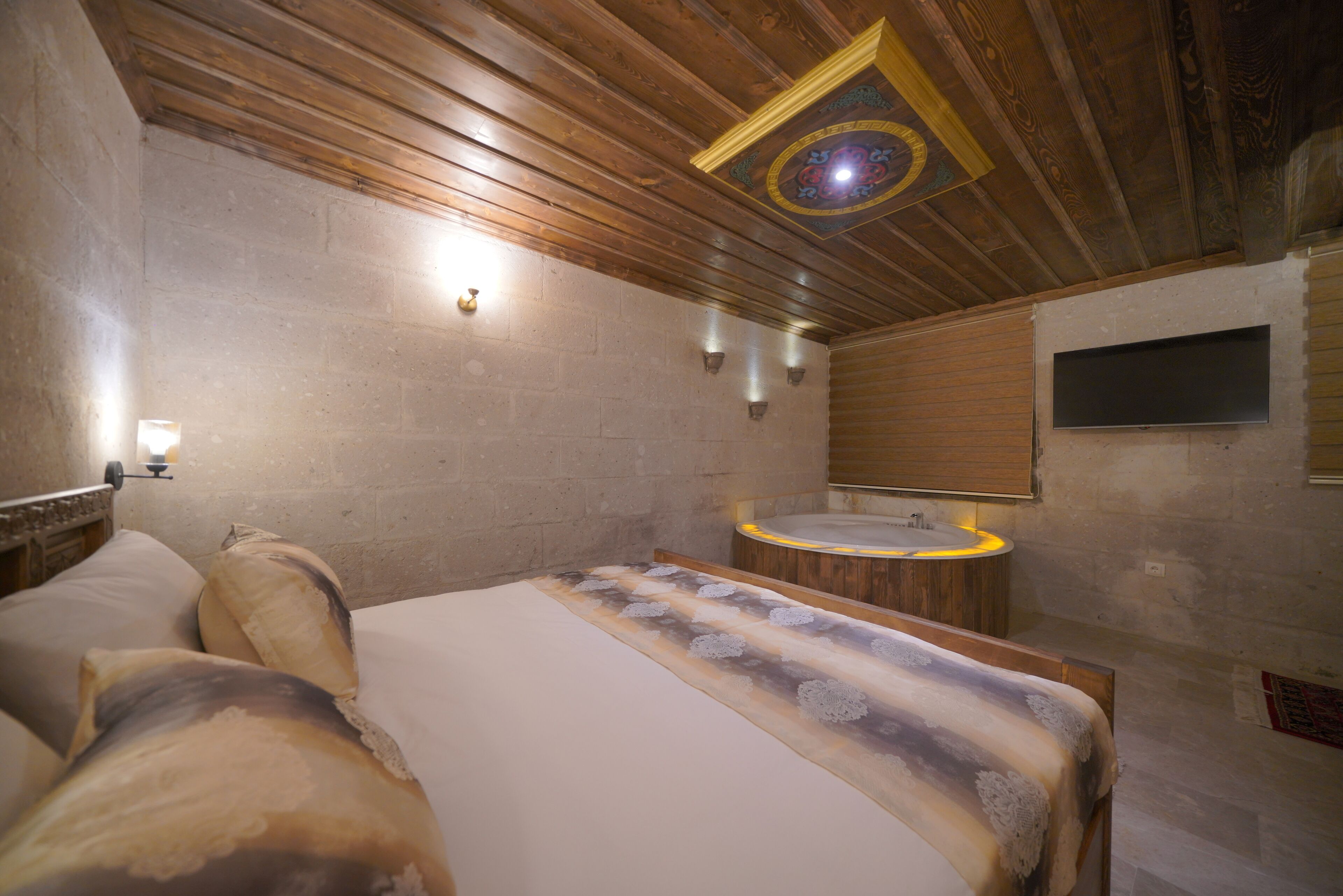 A bedroom with a wooden ceiling, a large bed with patterned bedding, a built-in bathtub, and a wall-mounted TV.