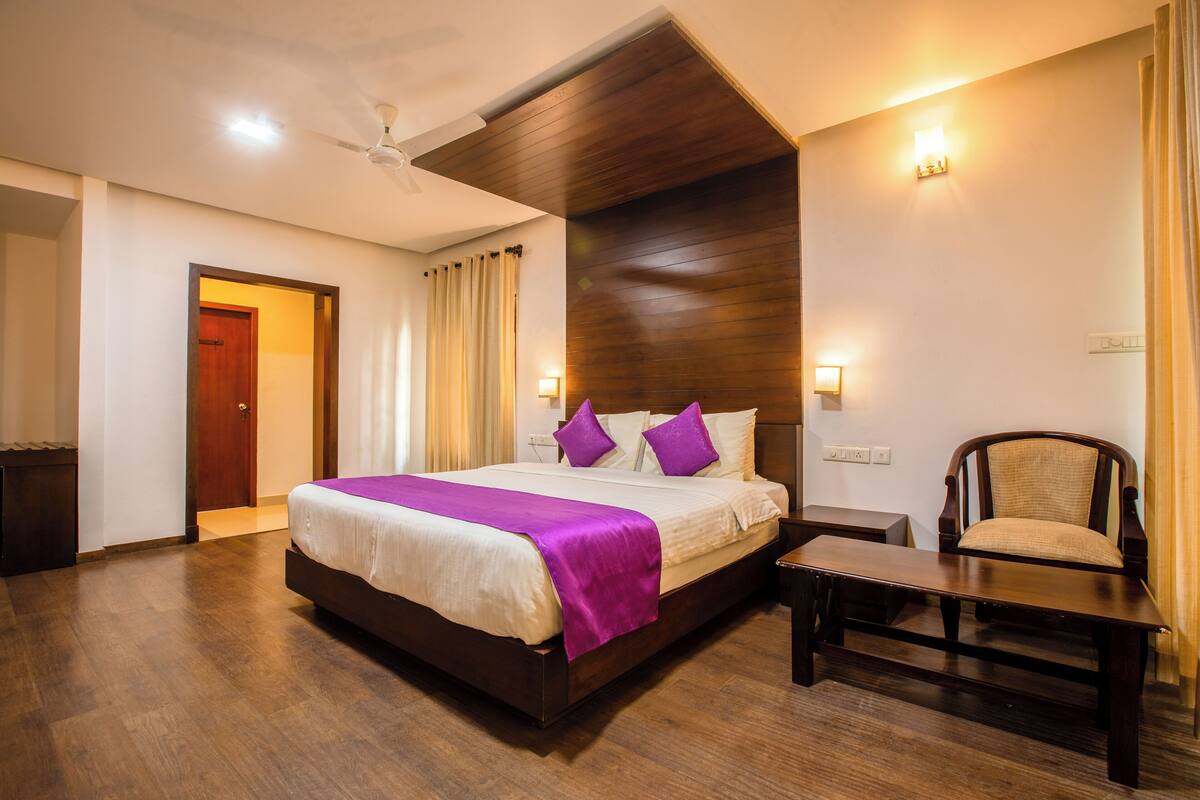A hotel room with a large bed, wooden floor, a ceiling fan, a nightstand, and a chair.