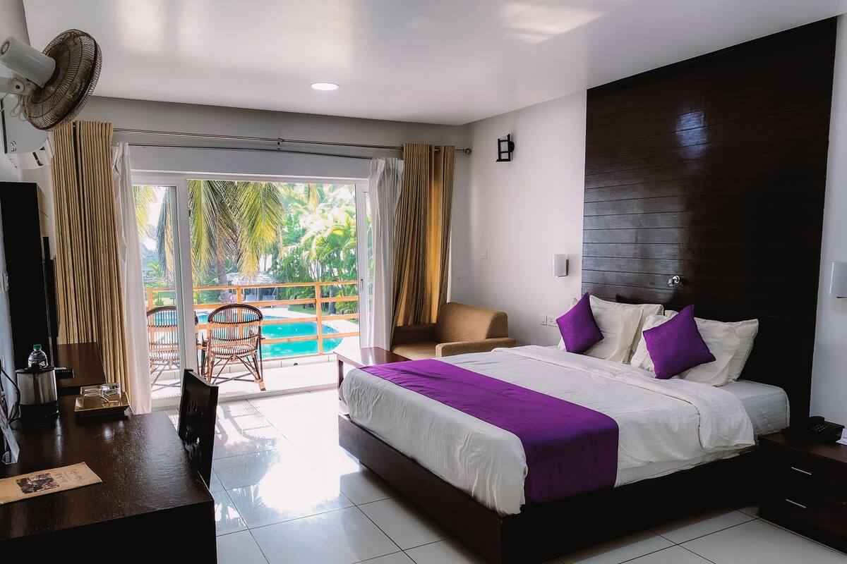 A modern hotel room with a large bed, a wooden headboard, a desk with a chair, and a view of a swimming pool.