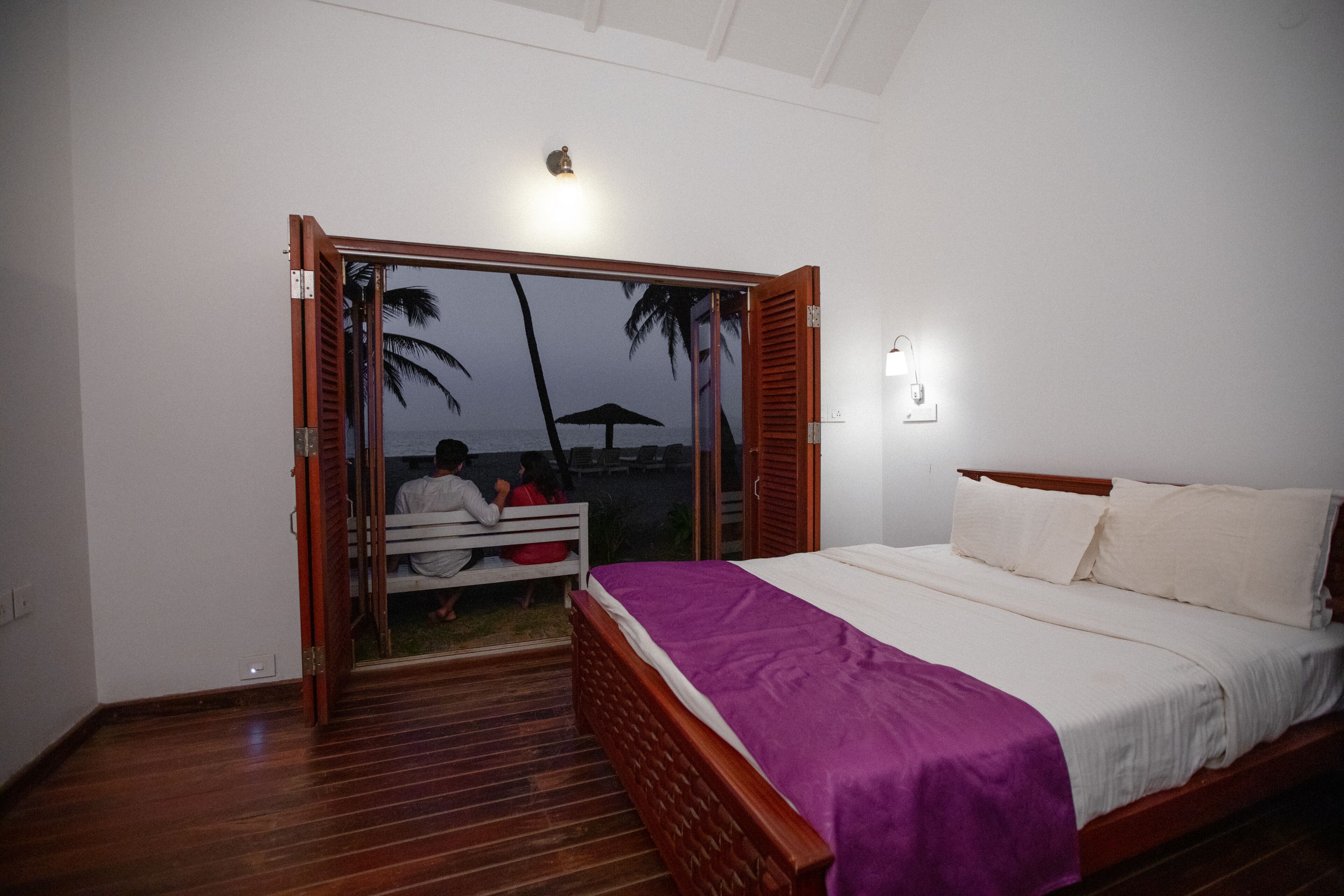 A bedroom with a wooden bed, a purple blanket, and a view of a beach with people sitting on a bench.