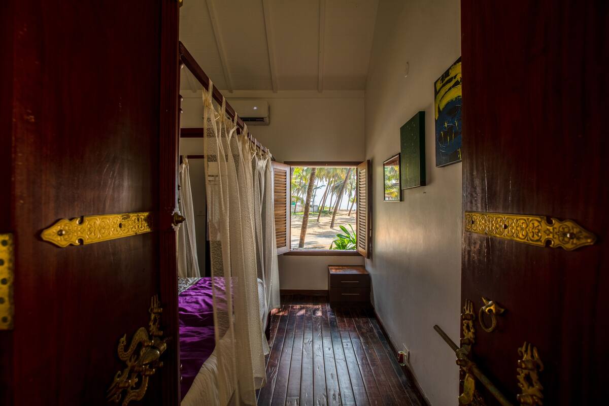 A narrow hallway with wooden flooring and ornate brass door handles. A window with wooden shutters and a view of palm trees is visible.