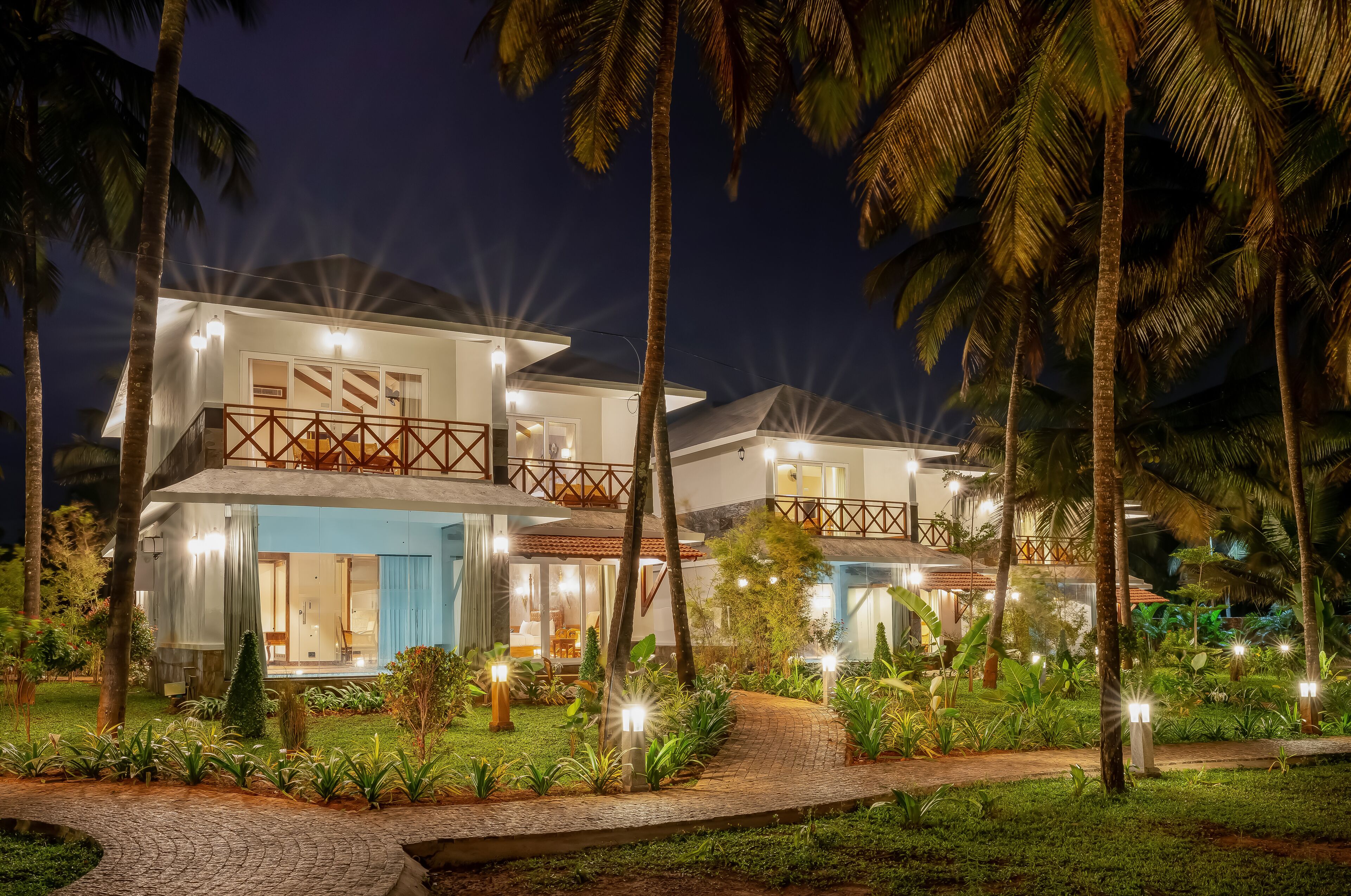 Un moderno resort de dos pisos con un espacio exterior bien iluminado, palmeras y un sendero pavimentado.