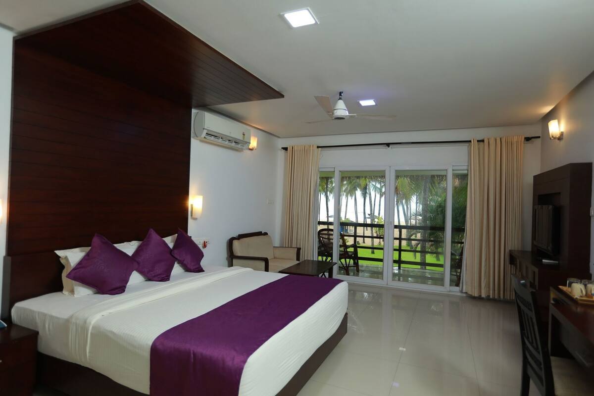 A hotel room with a bed, a TV, a ceiling fan, and a view of a beach.