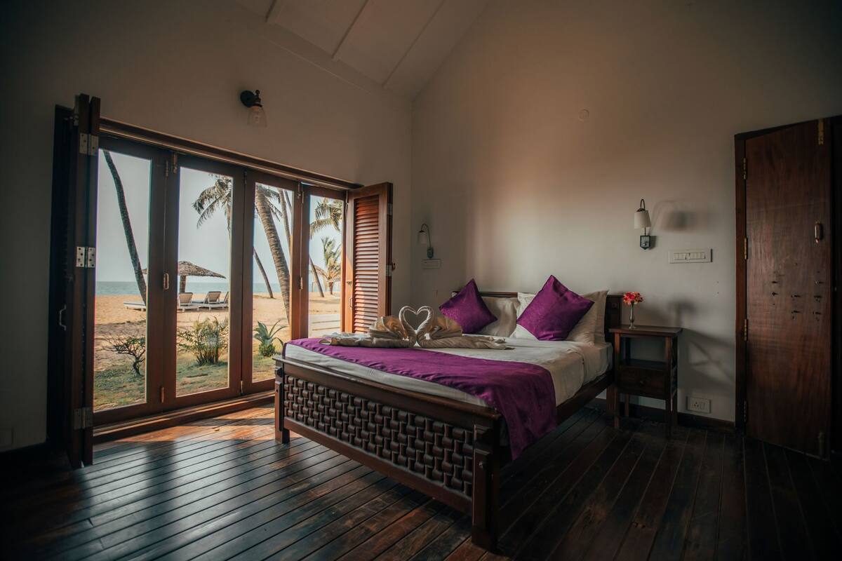 A bedroom with a bed, a nightstand, a wooden floor, a sliding glass door to a beach, and a view of palm trees.