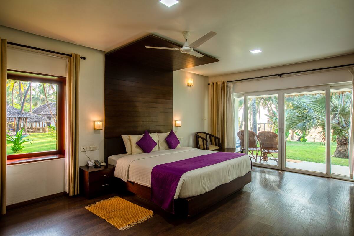 A spacious bedroom with a large bed, a ceiling fan, and a view of a lush garden through a sliding glass door.