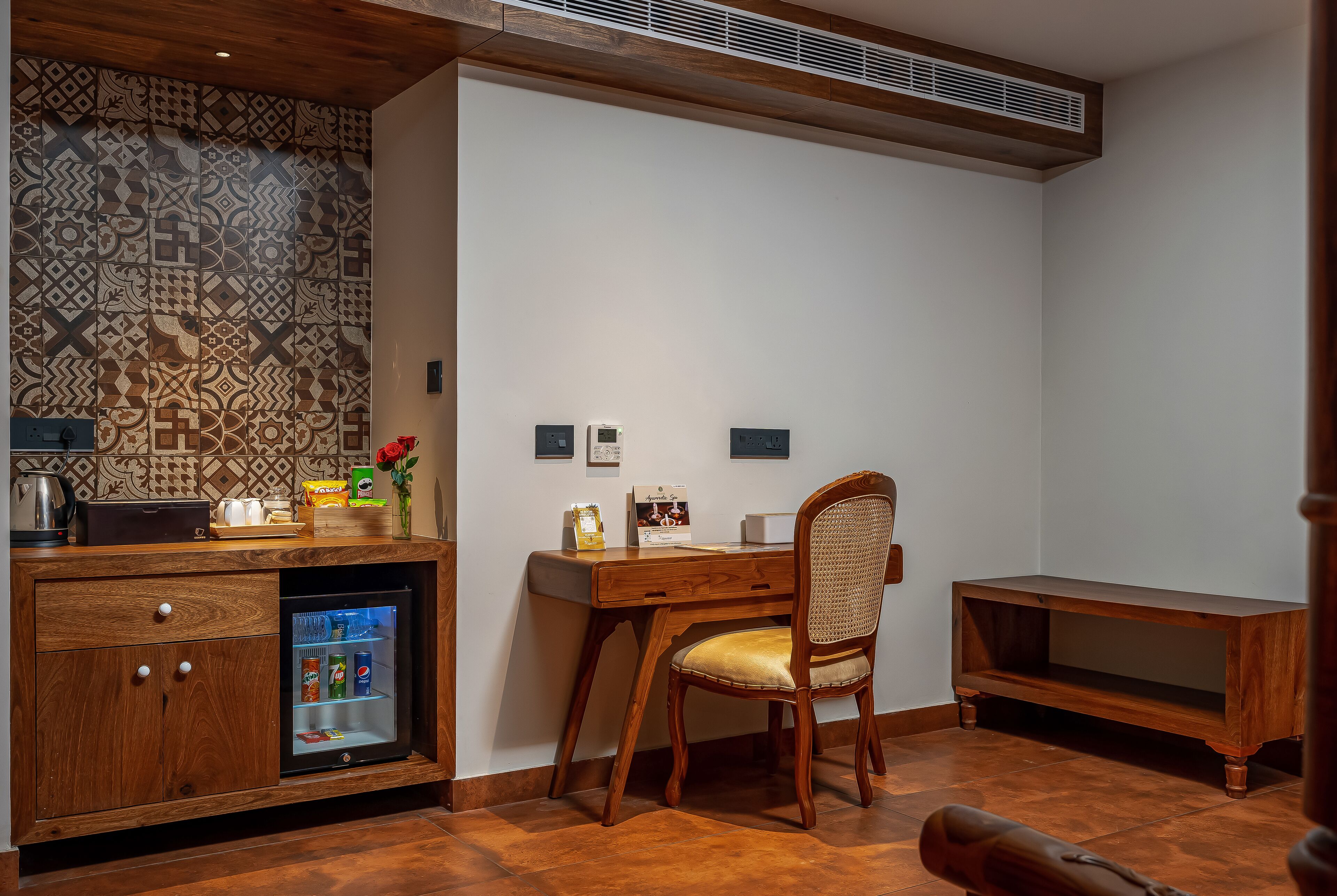 A compact room with a wooden desk, a chair, a built-in mini-fridge, and a small cabinet with drawers.