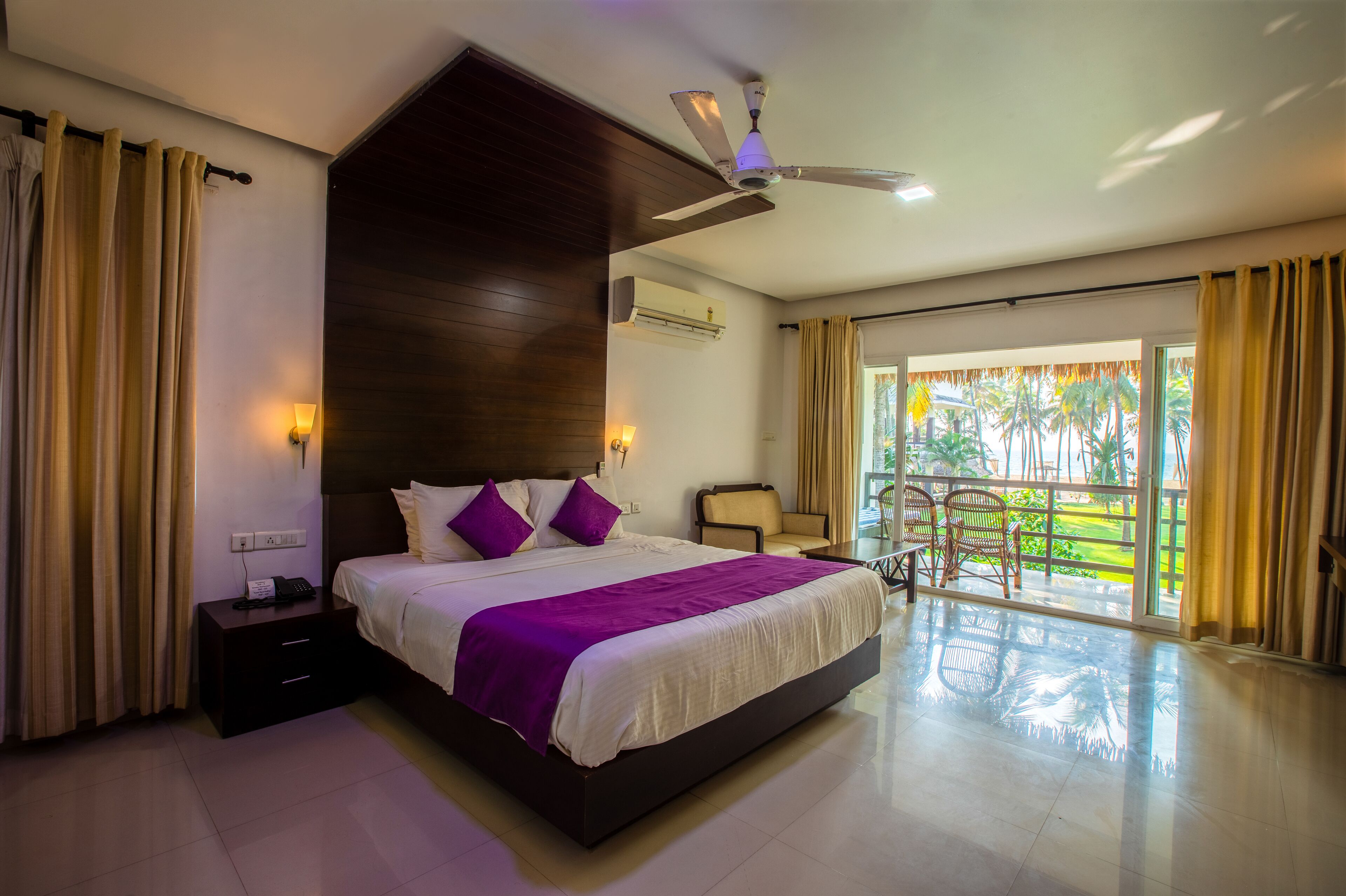 A modern hotel room with a large bed, a ceiling fan, a nightstand, a chair, and a view of palm trees through the balcony doors.