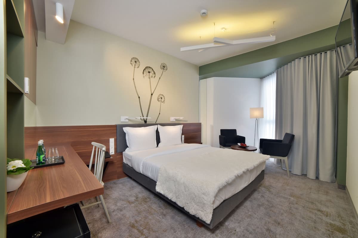 Quarto de hotel com cama, mesa de cabeceira, luminária, cadeira e vista para o exterior.