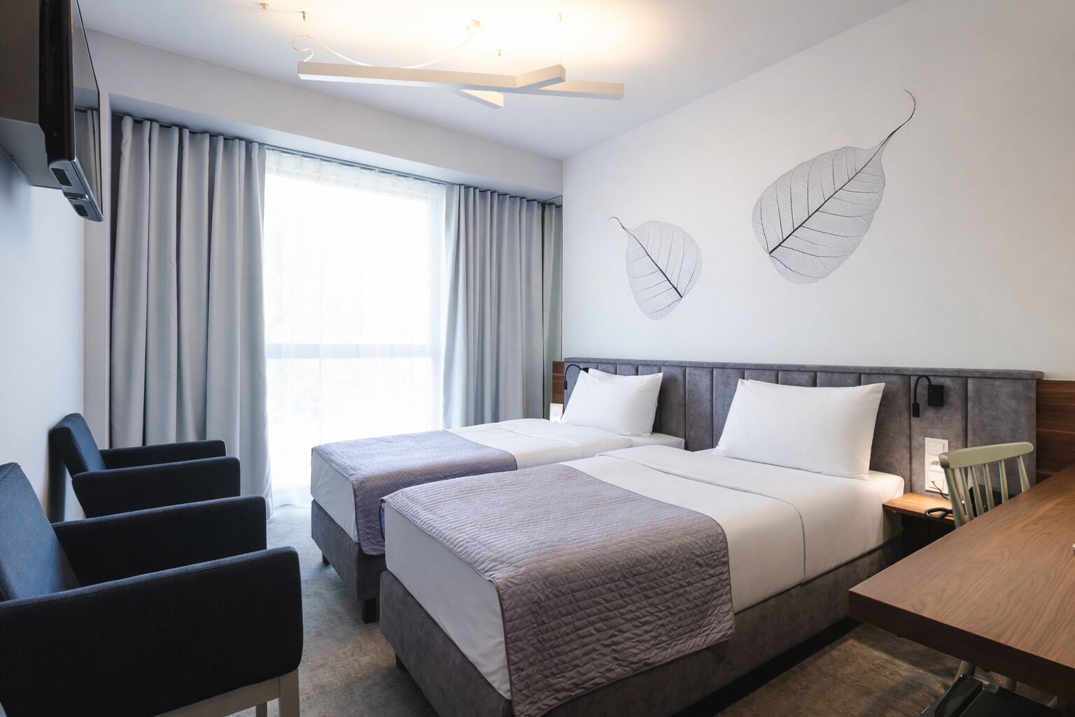 A hotel room with two beds, a desk, a chair, a TV, and a large window with curtains.