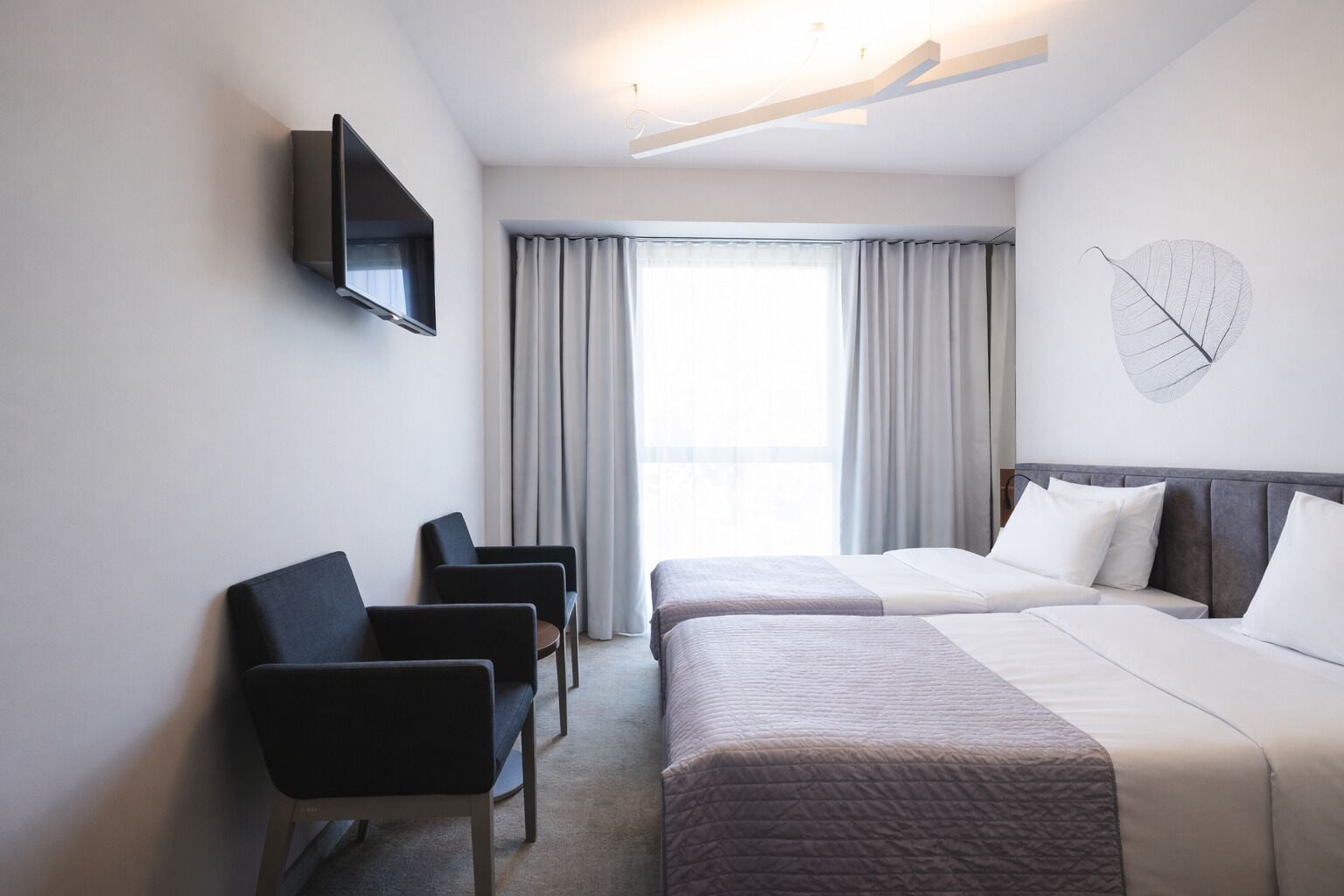 Habitación de hotel con dos camas, televisión, escritorio con sillas y una ventana grande con cortinas.