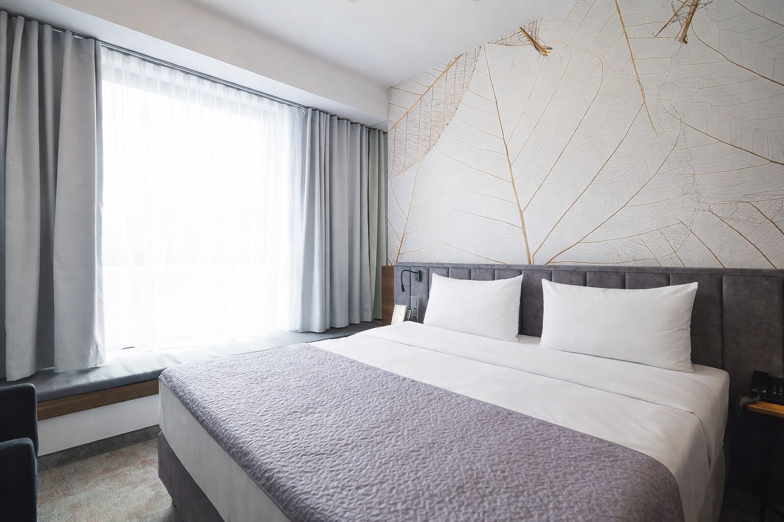 Habitación de hotel con una cama grande, cortinas grises, una lámpara de pared y un adorno decorativo en la pared con diseño de hojas.