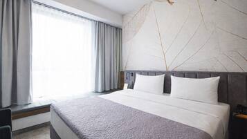 Habitación de hotel con una cama grande, cortinas grises, una lámpara de pared y un adorno decorativo en la pared con diseño de hojas.