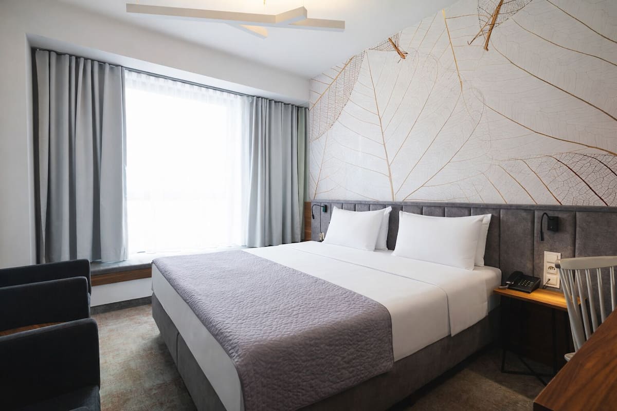 Quarto de hotel com cama grande, escrivaninha, cadeira e janela com cortinas.