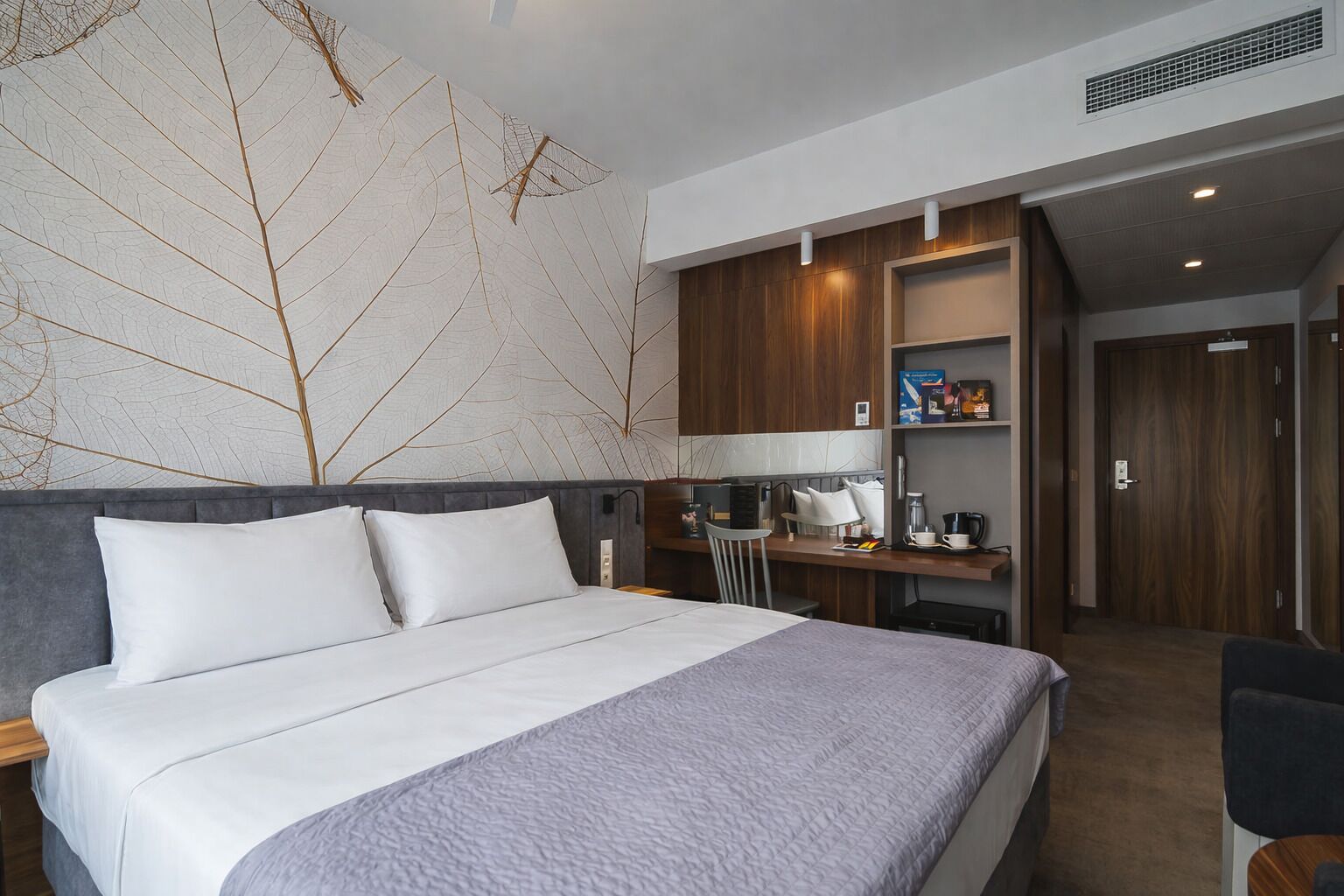 Una habitación de hotel con cama, escritorio, silla, cafetera y estante con artículos de tocador.