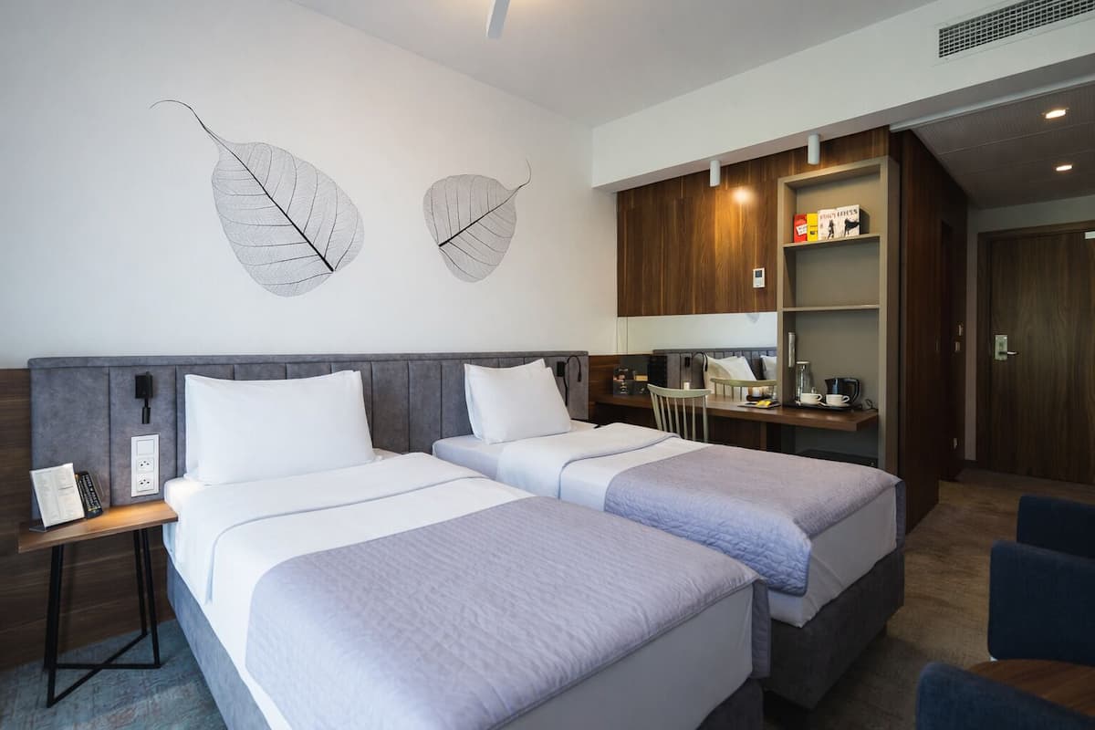 Quarto de hotel com duas camas, uma escrivaninha com cadeira, cozinha compacta e um guarda-roupa.