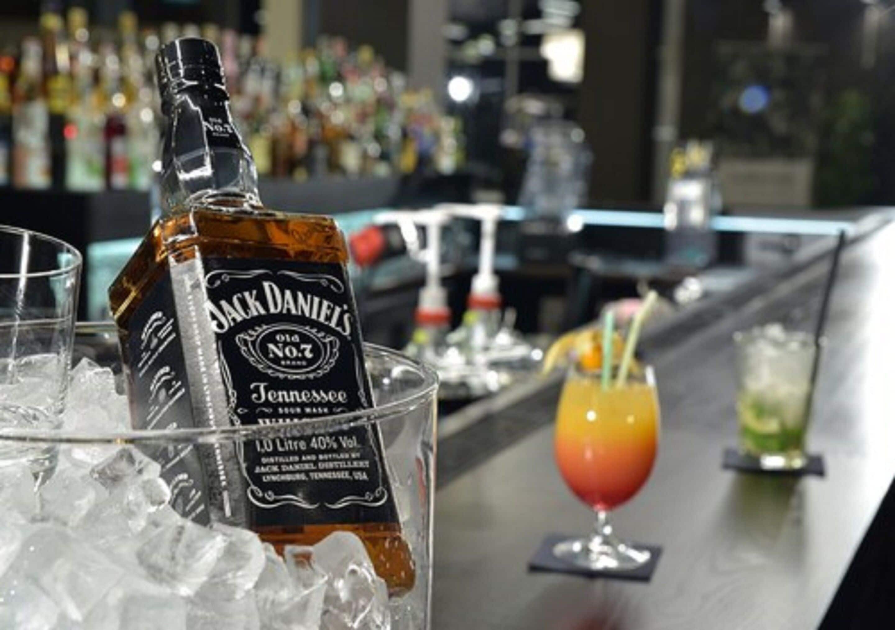 A bottle of Jack Daniel's whiskey on ice, a glass with a straw, and a cocktail with a lime garnish on a bar counter.