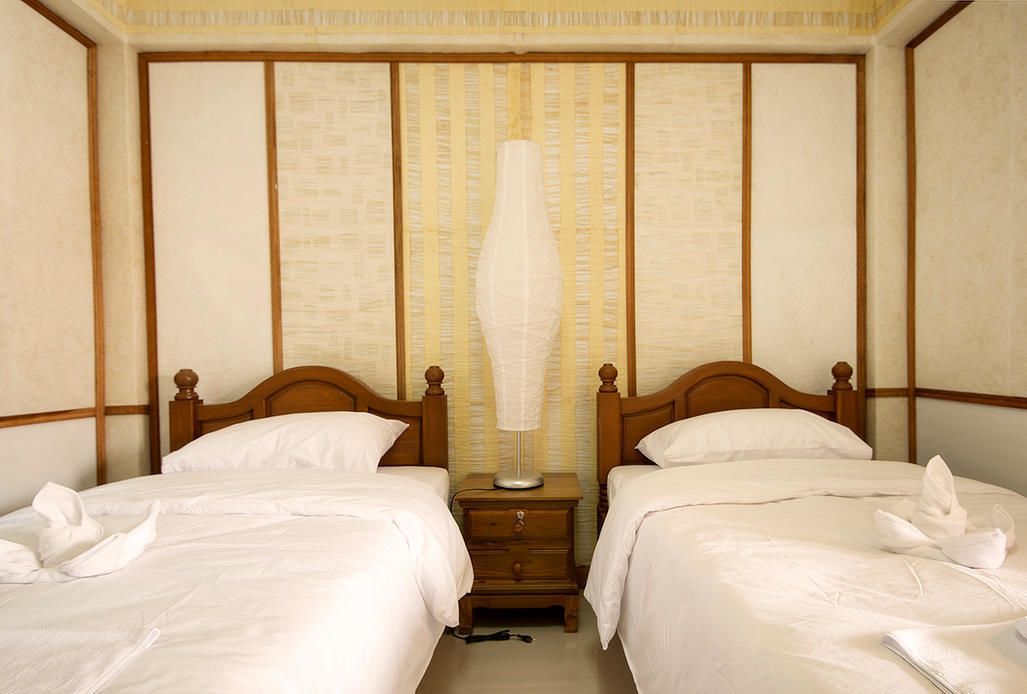 A hotel room with two beds, wooden headboards, a bedside table with a lamp, and a wall with vertical wooden panels.