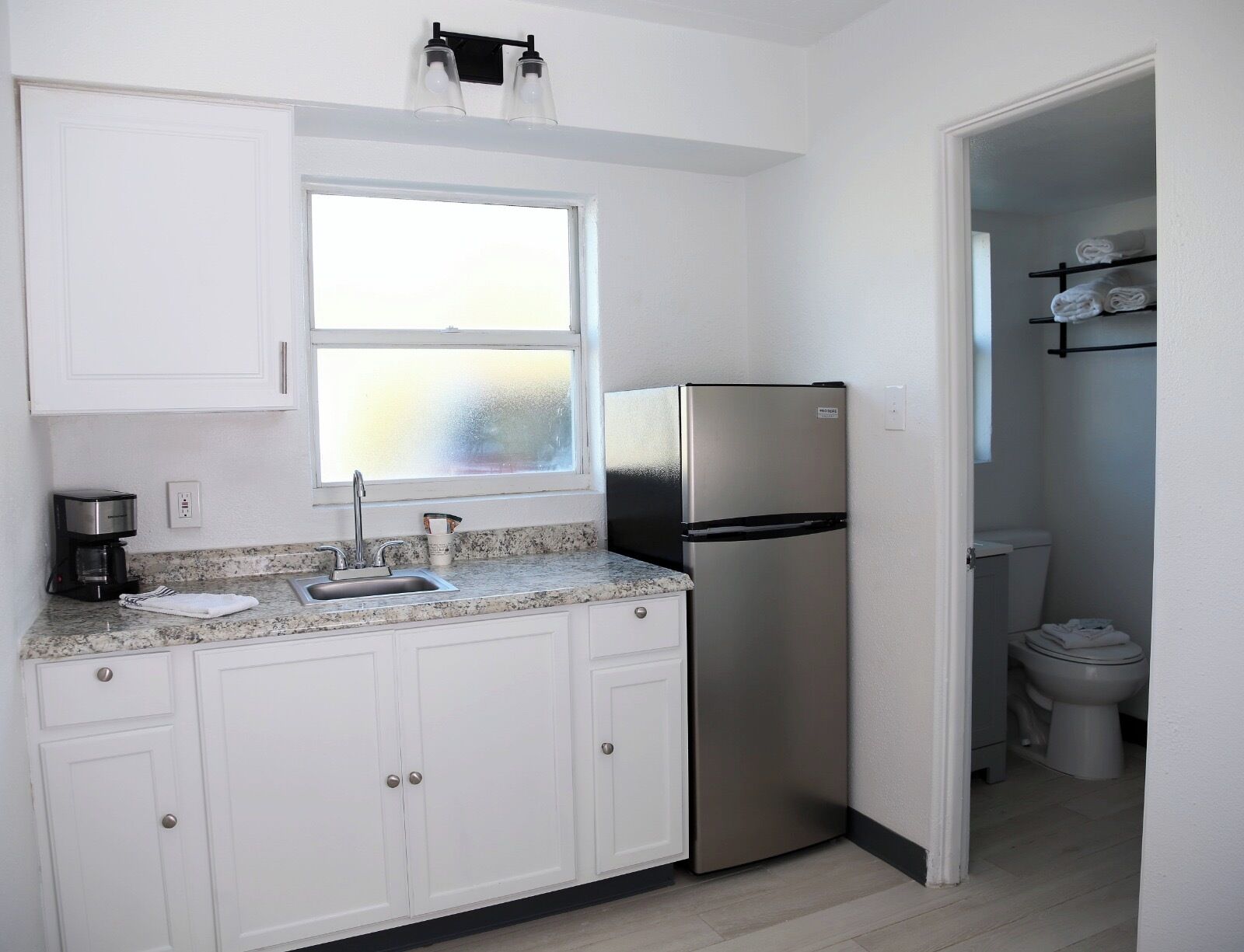 Cocina compacta con armarios blancos, un refrigerador de acero inoxidable, una cafetera y una ventana sobre el fregadero.