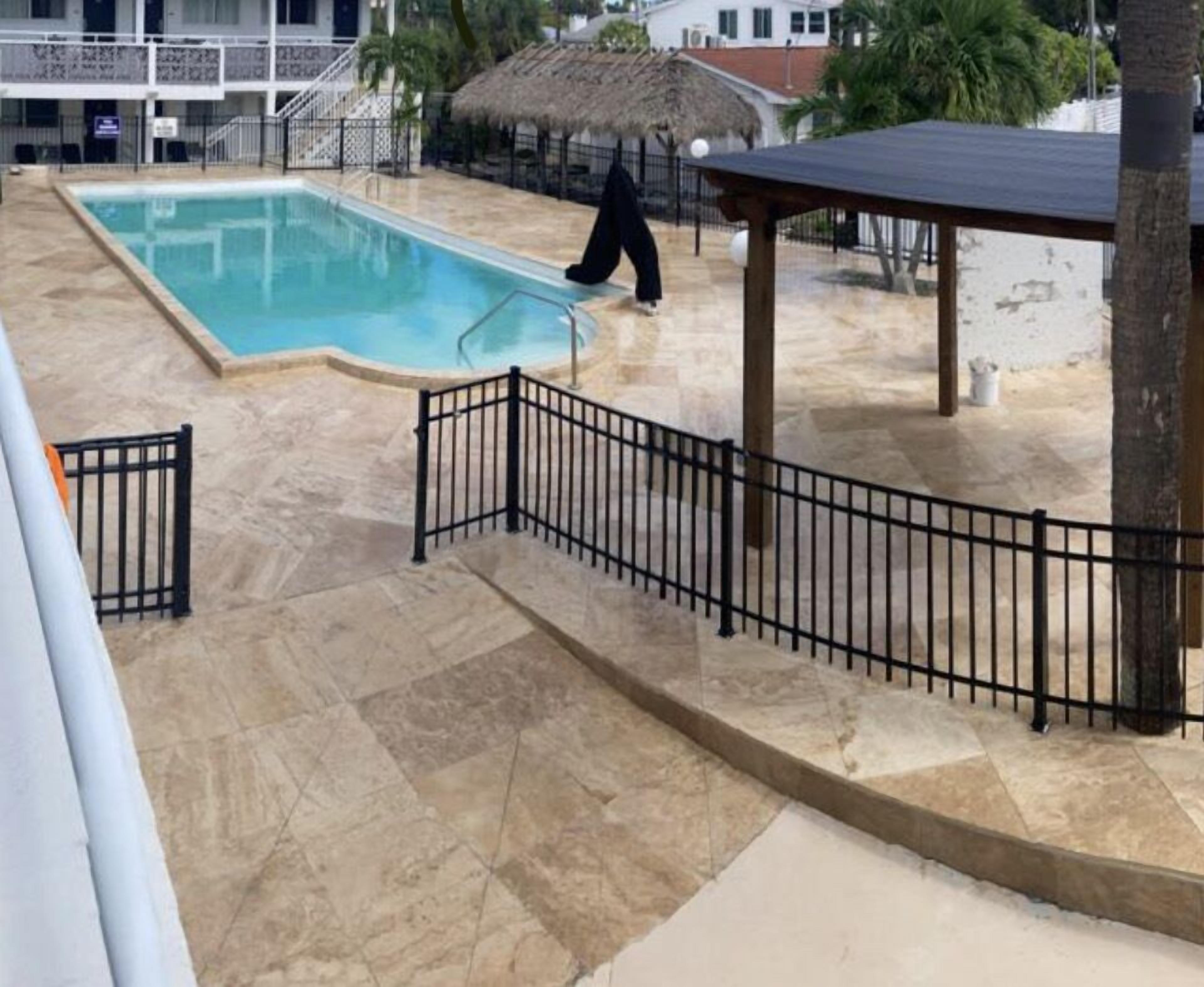 Una piscina con cercado de metal negro, un área de descanso cubierta y una estructura con techo de paja.