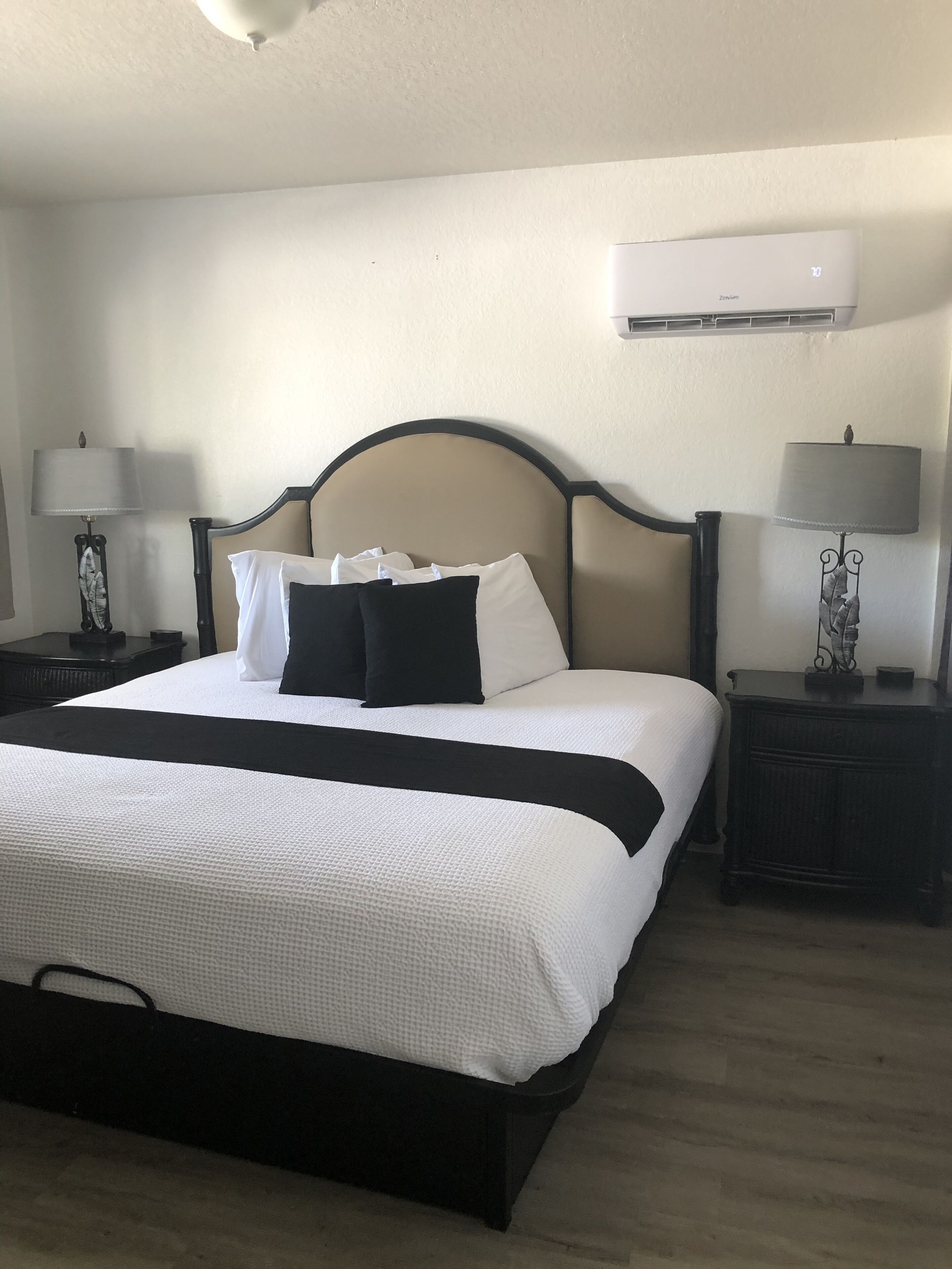 A neatly made bed with a black and white bedspread, two black pillows, and a white headboard. There are two bedside tables with lamps and a wall-mounted air conditioner above the bed.