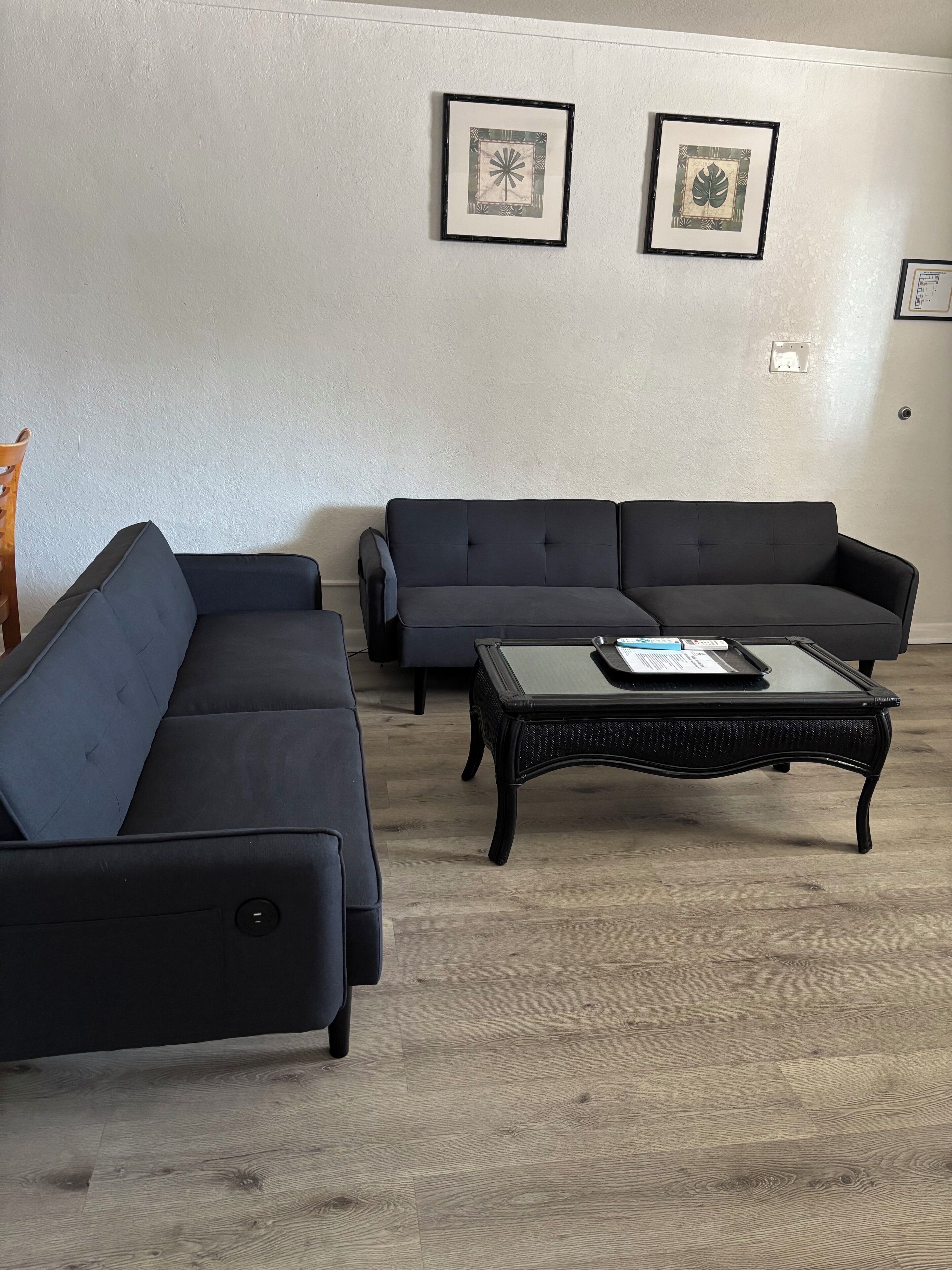 Una sala de estar con dos sofás oscuros, una mesa de centro de vidrio y dos obras de arte enmarcadas en la pared.