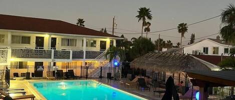 A swimming pool with lounge chairs and a covered seating area, surrounded by a fence and a building with balconies.