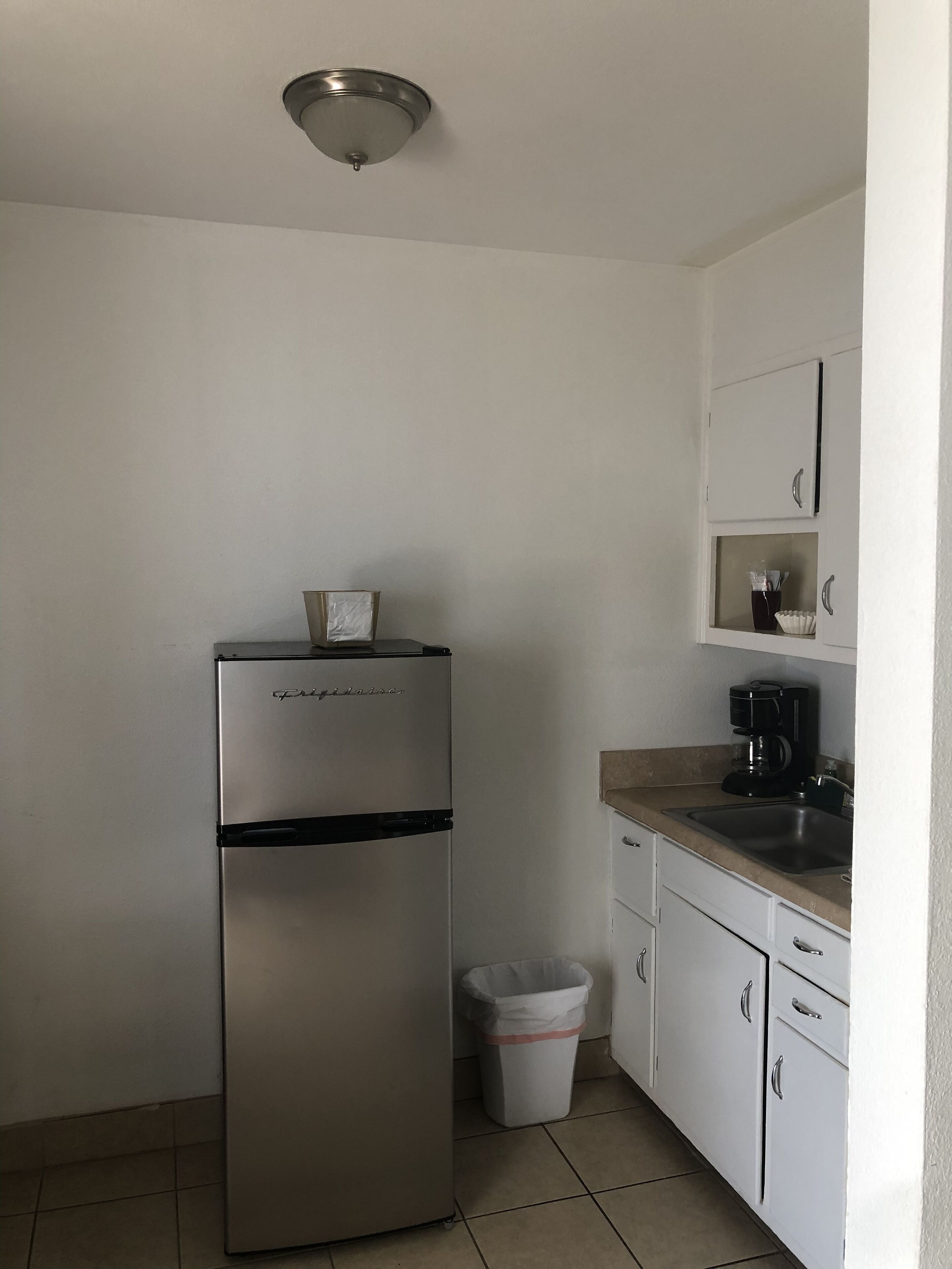A compact kitchen with a stainless steel refrigerator, a coffee maker, and a trash bin.