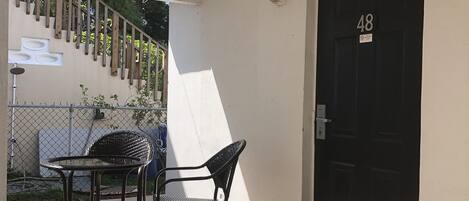 Un espace extérieur abrité, avec une table et deux chaises, une porte noire portant le numéro 48 et un mur blanc avec une fenêtre.