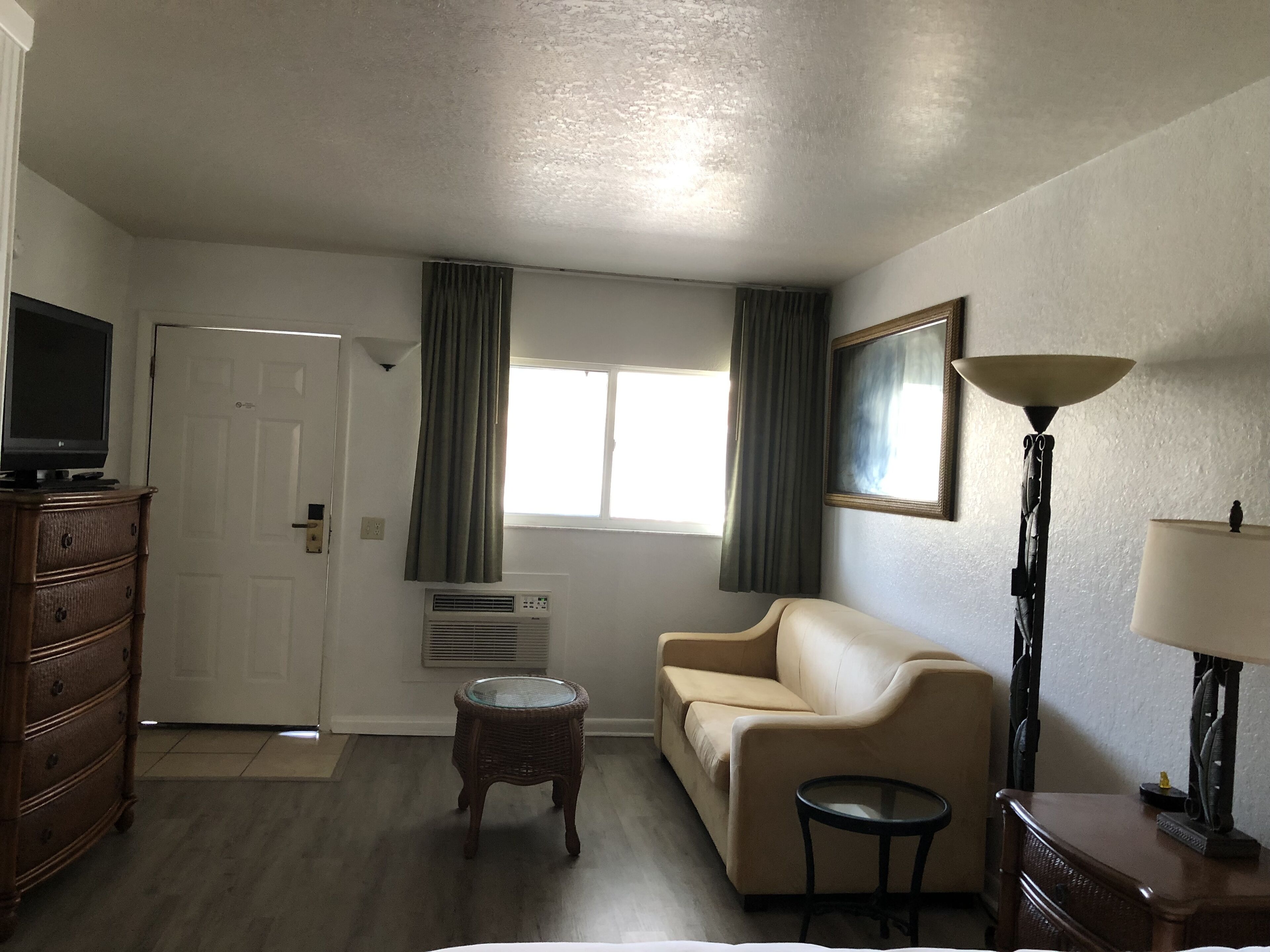 A living room with a beige sofa, a small round table, a wooden dresser, a television, a window with curtains, a floor lamp, and a wall-mounted picture frame.