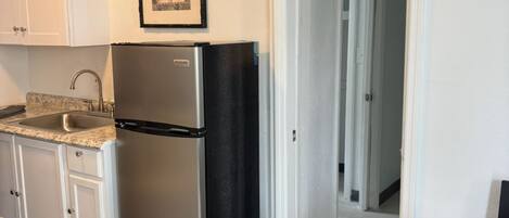 A compact kitchen with white cabinets, a stainless steel refrigerator, a granite countertop, and a framed picture above the fridge.