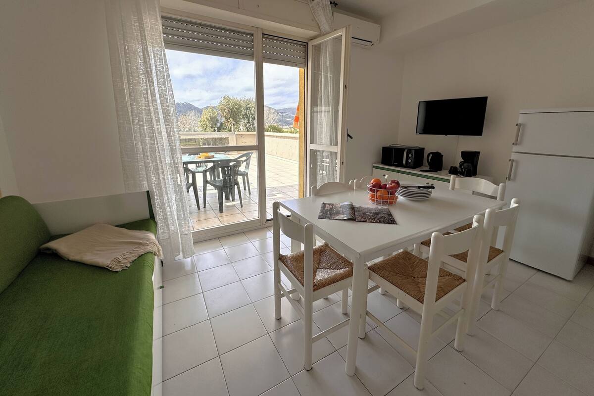 Una cocina limpia y moderna con una mesa de comedor, sillas y un refrigerador. Un televisor está montado en la pared, y hay una puerta corrediza de vidrio que da a un área exterior con una mesa y sillas.