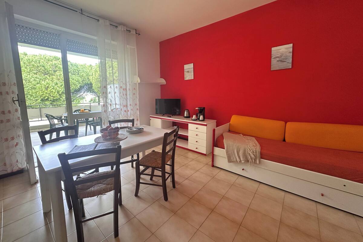 Un comedor con una mesa, sillas y un televisor. Hay un sofá con un cojín naranja y una pared roja.