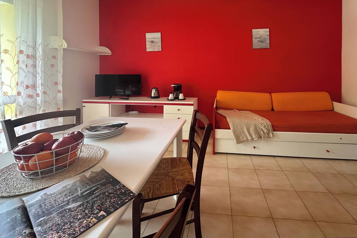 Una habitación con pared roja, cama con ropa de cama naranja, escritorio con televisor, cafetera y zona de comedor con una cesta de naranjas y una revista.