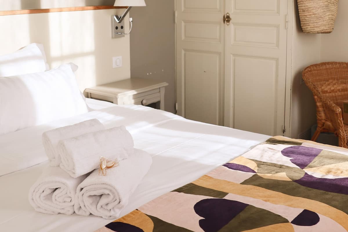 A hotel room with a neatly made bed, a large wardrobe, a bedside table with a lamp, and a wicker chair.