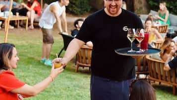 Een persoon serveert drankjes aan gasten die zitten aan buitentafels met parasols.