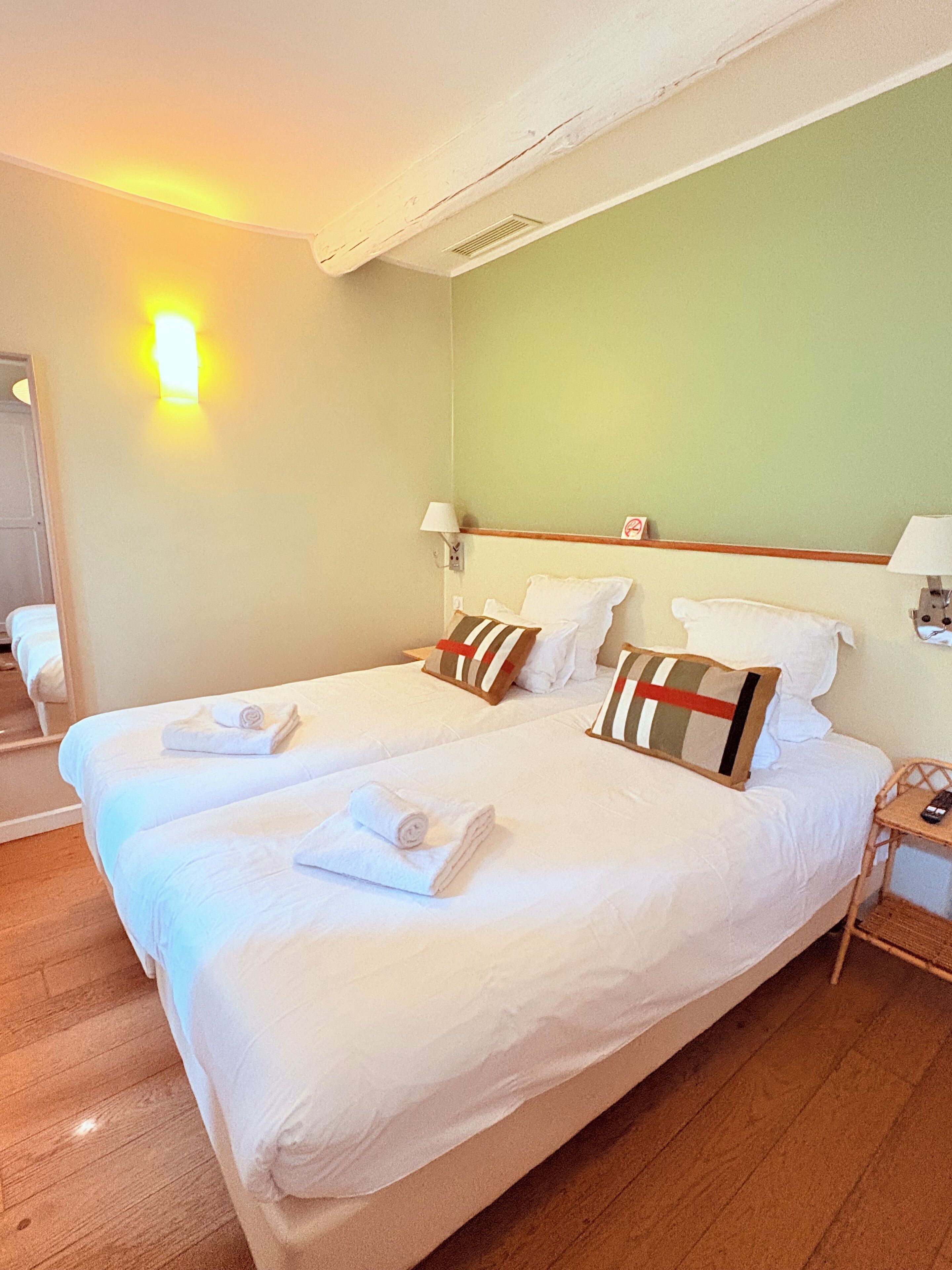 A double bed with white linens and striped pillows, a bedside table with a lamp, and a wooden floor.