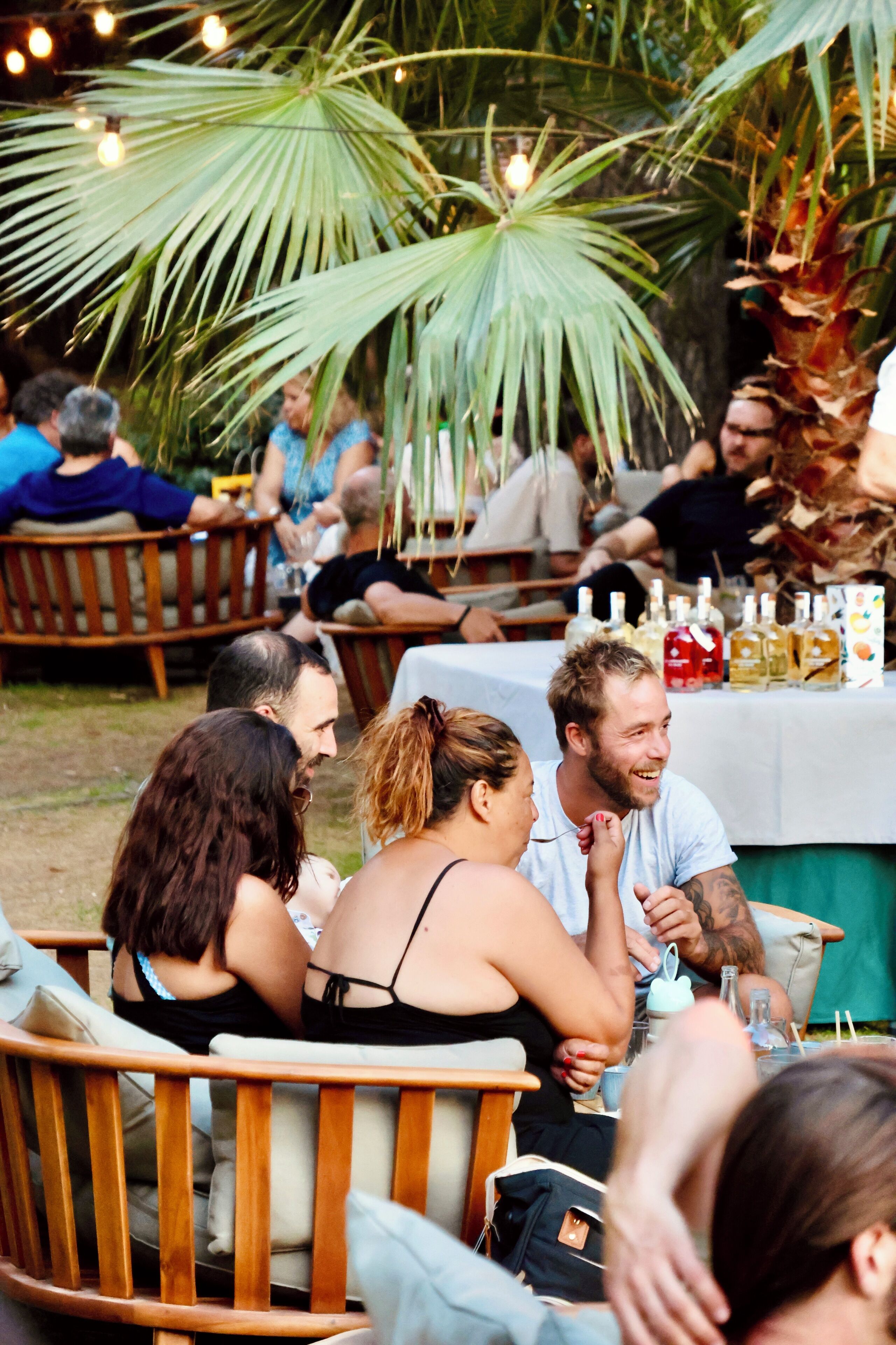 A group of people seated at an outdoor table with a white tablecloth, surrounded by wooden chairs and palm trees.
