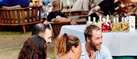 Un groupe de personnes est assis autour d’une table en plein air, recouverte d’une nappe blanche, et entouré de chaises en bois et de palmiers.