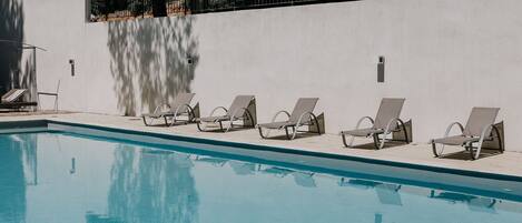 A swimming pool with six sun loungers aligned along the edge, a white wall, and a metal fence in the background.