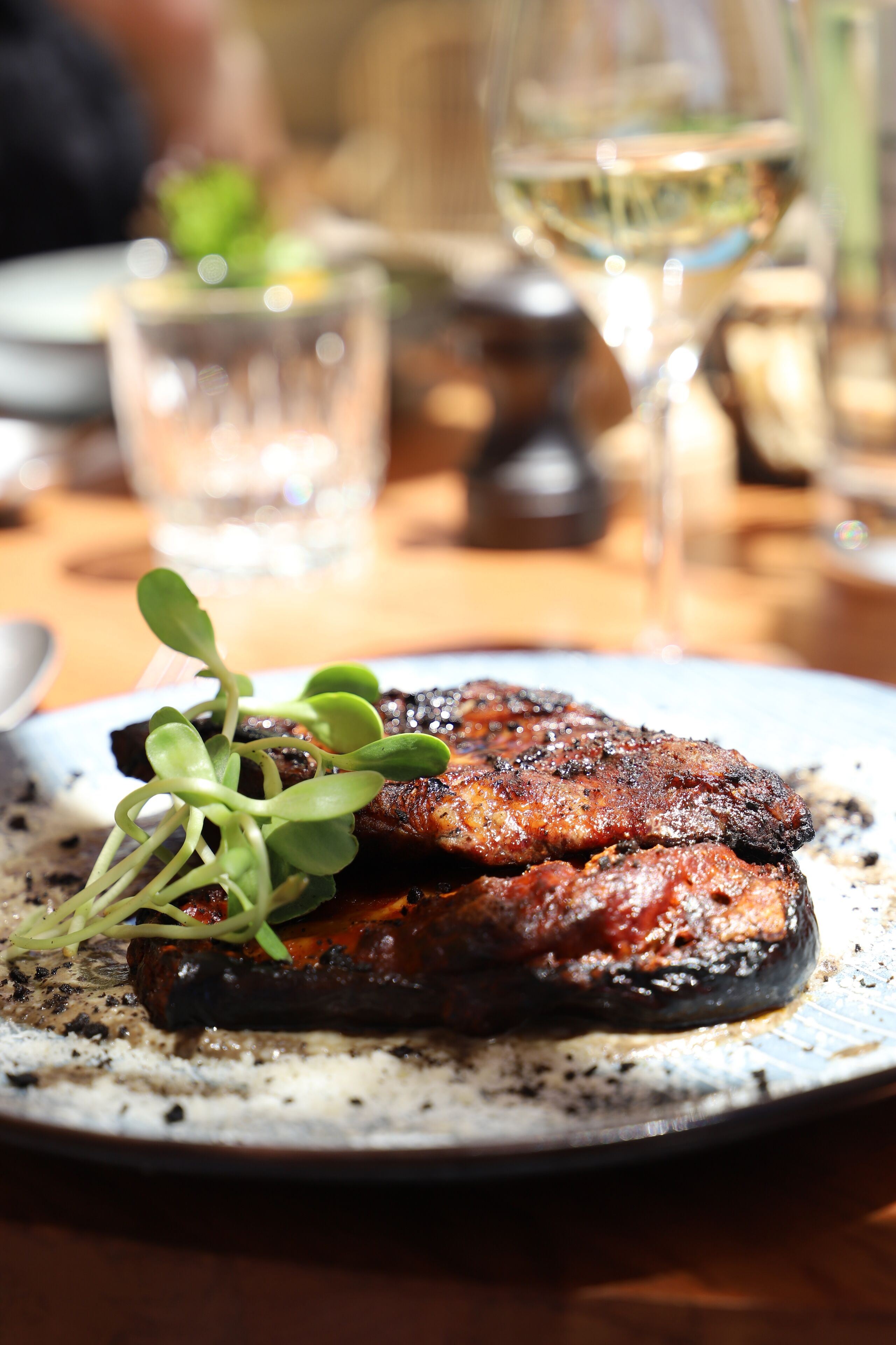 A plate with grilled meat and greens, a glass of white wine, and a blurred background.