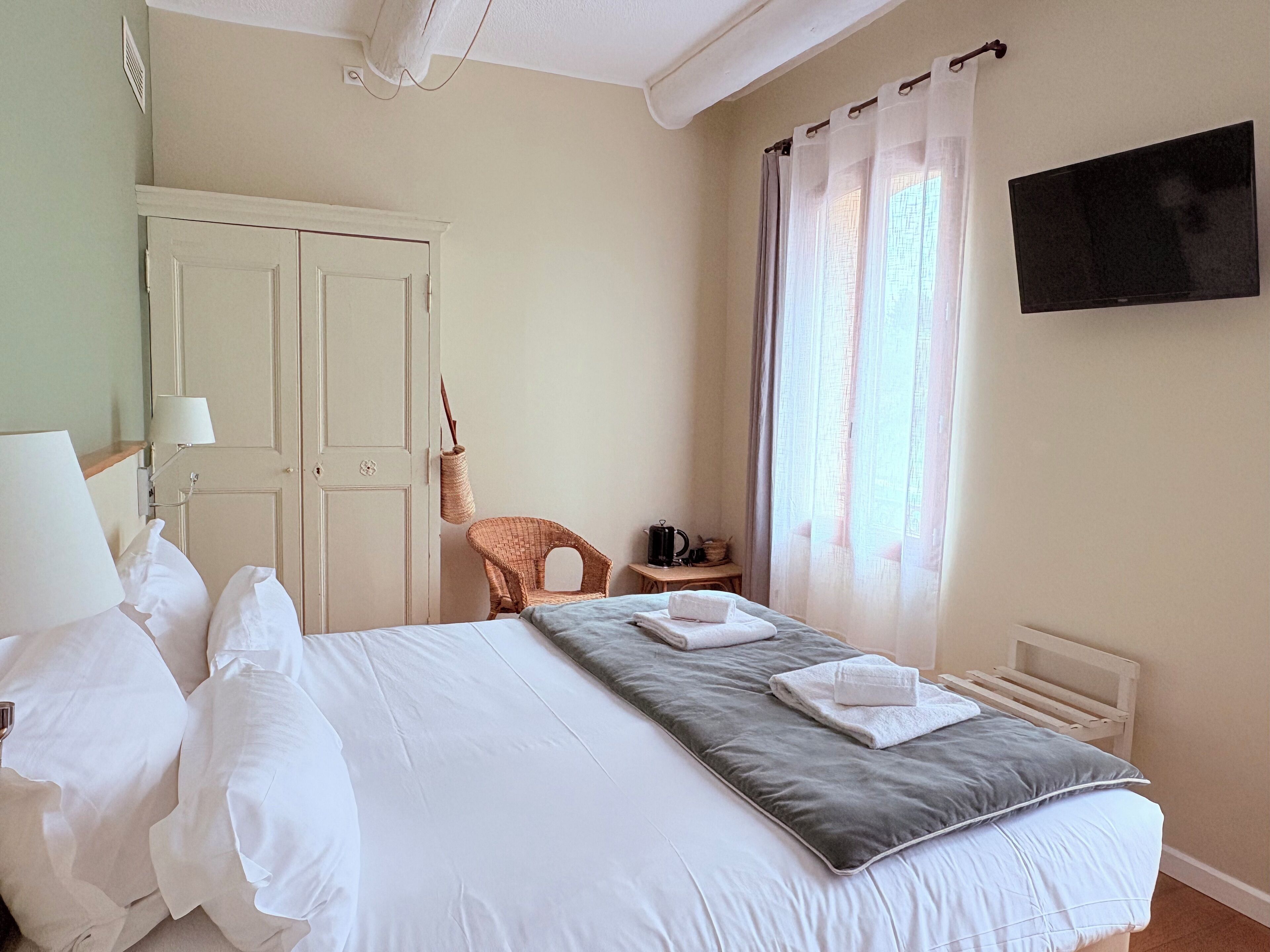 A hotel room with a large bed, a chair, a small table with a kettle, and a TV on the wall.