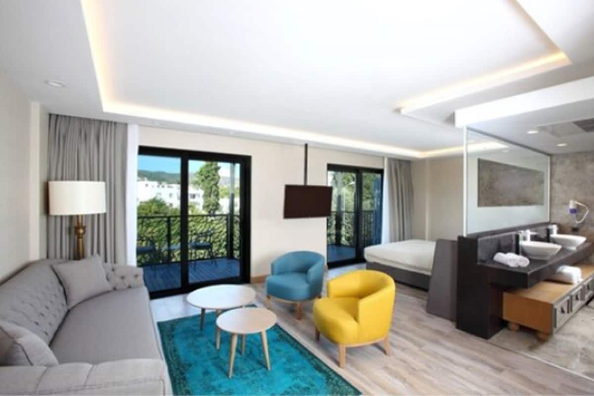 A modern hotel room with a sofa, two armchairs, a small round table, a large window with a balcony, a TV, a bathroom with a sink and mirror, and a blue rug.