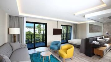 A modern hotel room with a sofa, two armchairs, a small round table, a large window with a balcony, a TV, a bathroom with a sink and mirror, and a blue rug.