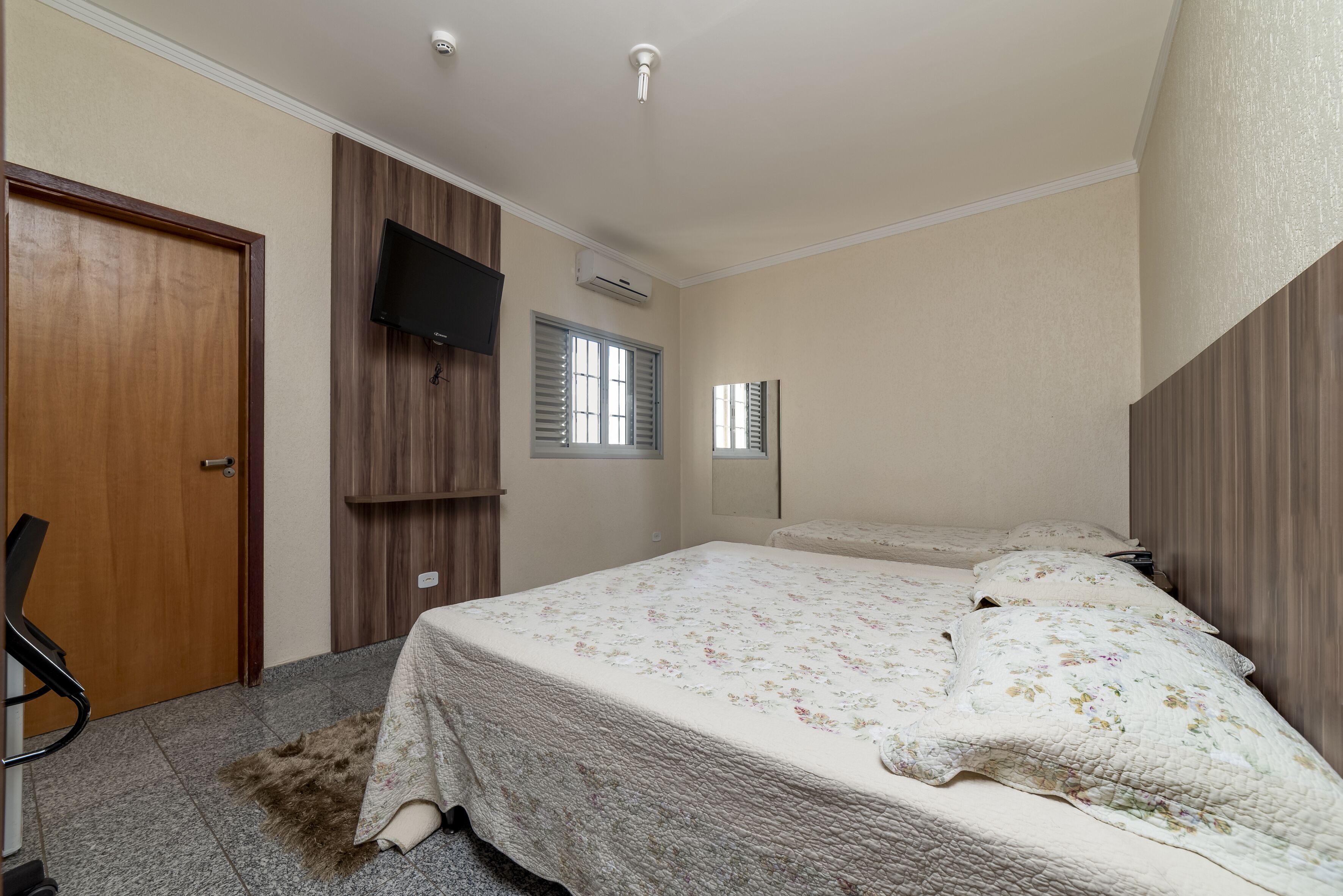 A bedroom with a bed, a TV mounted on the wall, an air conditioning unit, and a door.
