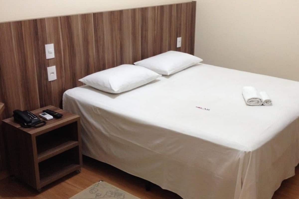Una cama bien hecha con sábanas blancas, una mesita de noche con teléfono y controles remotos, y un cabecero de madera.