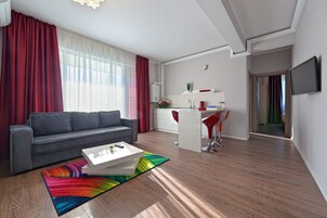 A modern living room with a grey sofa, a colorful rug, and a white coffee table. There is a kitchen area with white cabinets, a red chair, and a TV mounted on the wall.