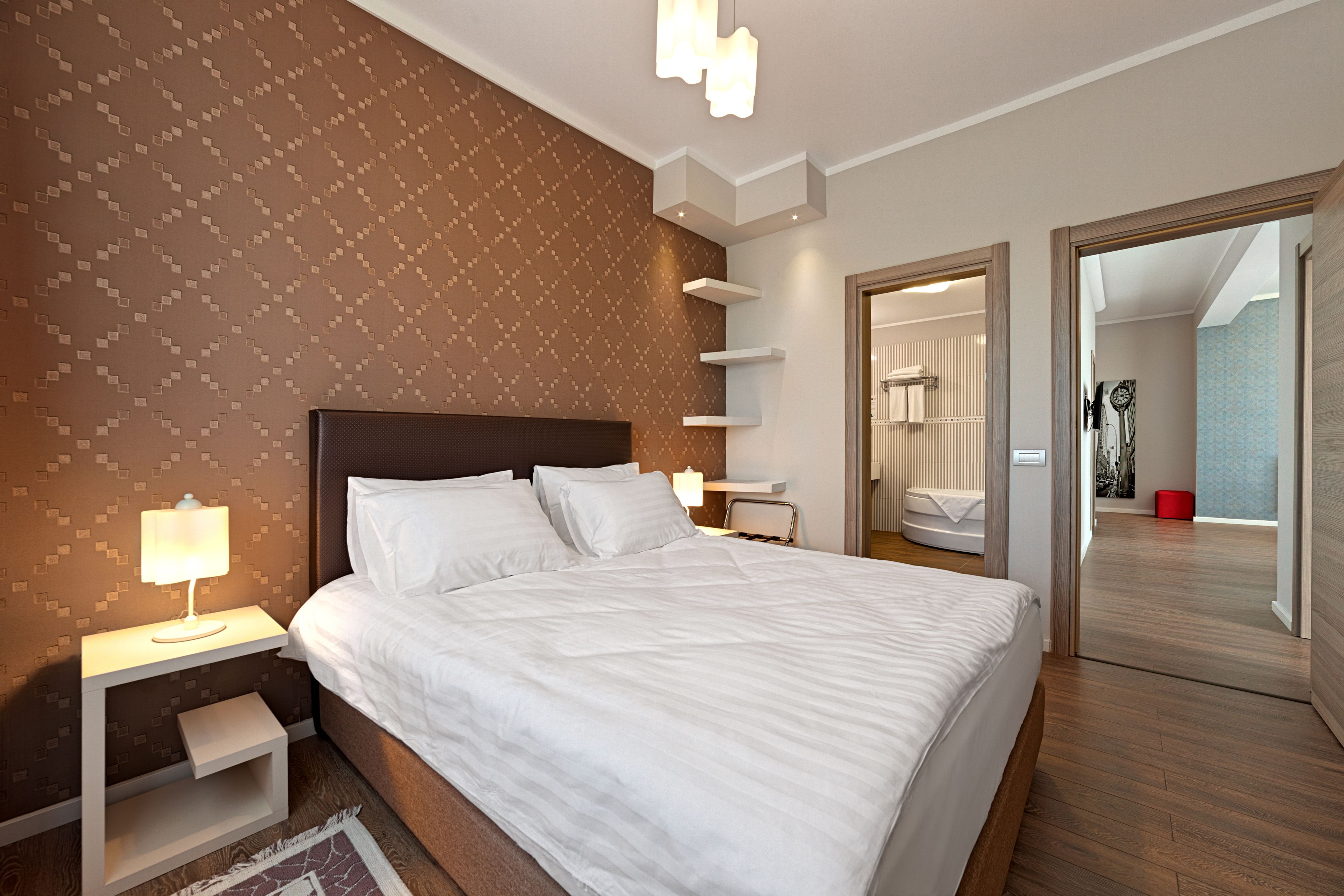 Quarto de hotel moderno com uma cama grande, mesa de cabeceira com abajur, prateleira embutida e vista para o banheiro.