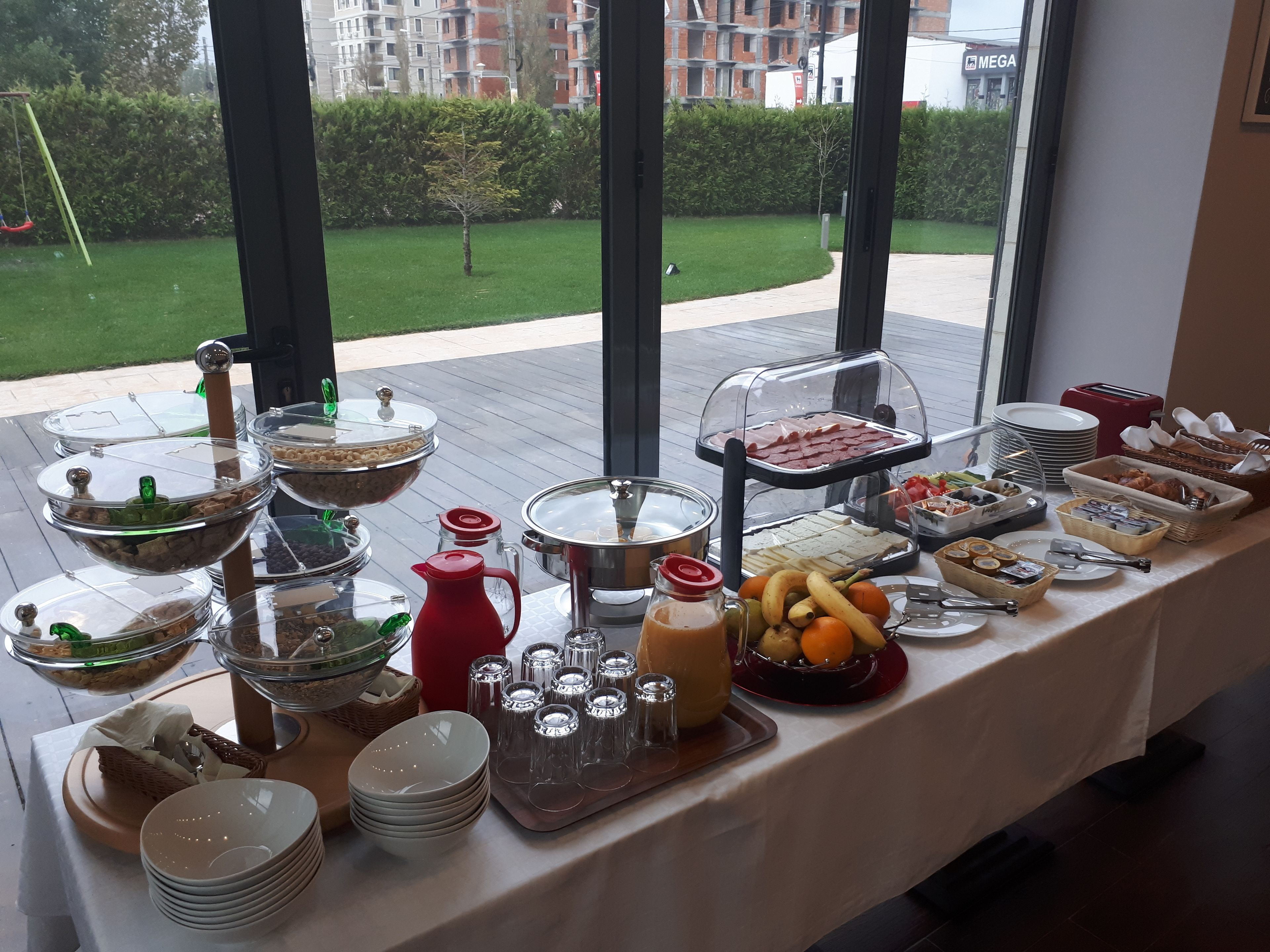 Une table de buffet garnie de divers plats et accompagnements, placée devant une fenêtre donnant sur un quartier résidentiel.