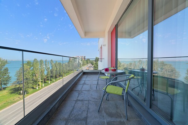 A balcony with a glass railing, a table with chairs, and a view of the sea and buildings.