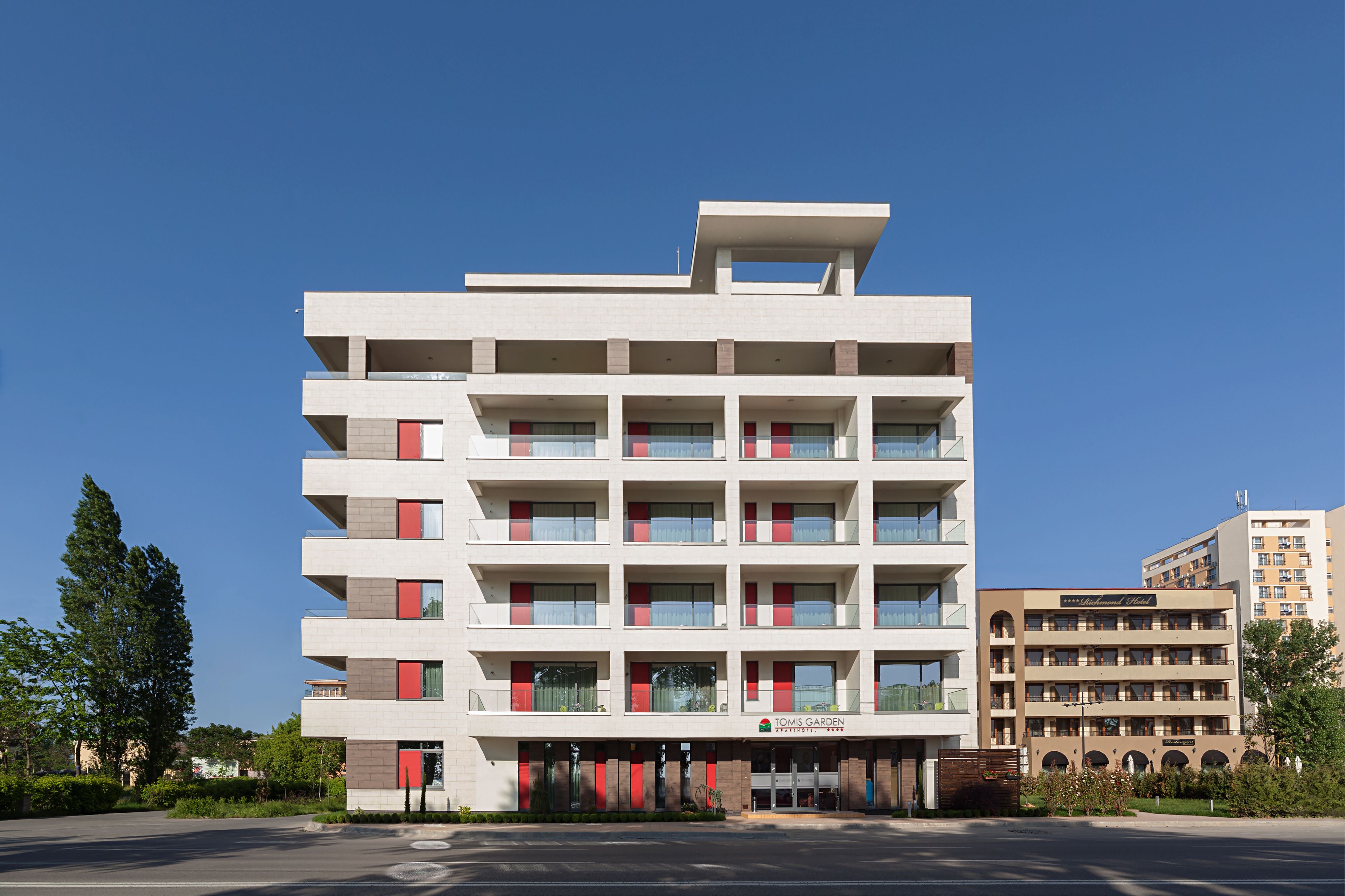 A multi-story hotel with a modern design, featuring balconies on each floor, a central entrance, and a parking lot in the foreground.